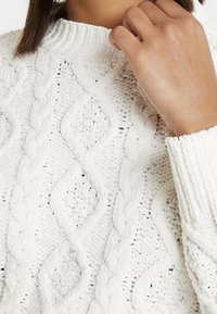 Pull en tricot blanc avec un motif en treillis et des formes de diamant, col montant et manches à bords côtes. La laine épaisse et texturée offre de la chaleur.