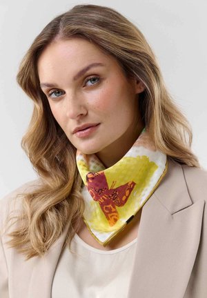 Woman with long wavy blonde hair wears beige blazer and white top, accessorized with a yellow scarf featuring an orange butterfly design.