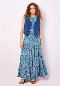 Denimweste über einem blumigen Maxikleid mit blauen, rosa und grünen Mustern; Rüschensleeves und gestuftem Rock; braune Sandalen.