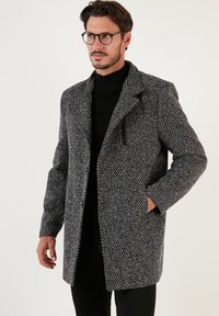Abrigo de tweed gris y negro con cuello, bolsillo frontal tipo solapa y patrón texturizado, llevado sobre un suéter de cuello alto negro.