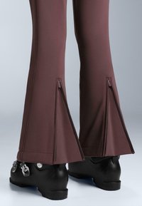 Donkerbruine skibroek met zijritsen bij de enkels; gemaakt van een rekbare stof. Zwarte skischoenen met zilverkleurige details zichtbaar eronder.