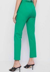 Groene broek met een op maat gemaakte pasvorm, enkellang, gladde textuur en riemlussen; gedragen met zwarte hoge muiltjes.