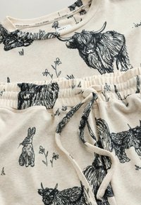 Beige bomuldsoutfit med en top og shorts. Stoffet viser sorte illustrationer af dyr og blomster, med elastisk talje.