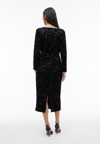 Sieviete ar garām tumšām matiem, ģērbta melnā mirdzošā midi kleitā ar garām piedurknēm un atvērumu aizmugurē, kopā ar melnām augstpapēžu kurpēm.