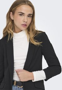 Črn krojen blazer preko bele turtleneck majice, ki se ponaša z gladko teksturo in odrezanimi reverji. Pas ima zlat poudarek.