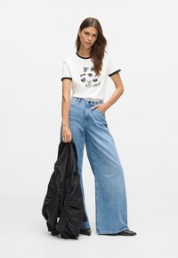 T-shirt de algodão branco com colarinho preto e estampa gráfica. Jeans azuis de cintura alta e perna larga. Casaco puffer preto segurado com uma mão.