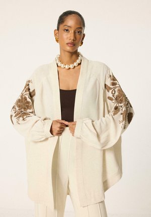 Femme portant une veste kimono crème avec broderie florale marron sur les manches, collier de perles, boucles d'oreilles en or et pantalon crème sur un haut foncé.