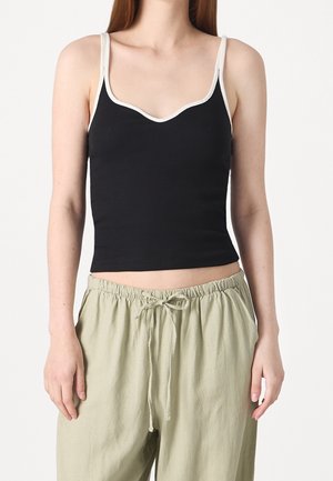 Vrouw draagt een zwarte mouwloze cropped top met witte biezen en lichtbeige broek met een trekkoord tegen een effen achtergrond.
