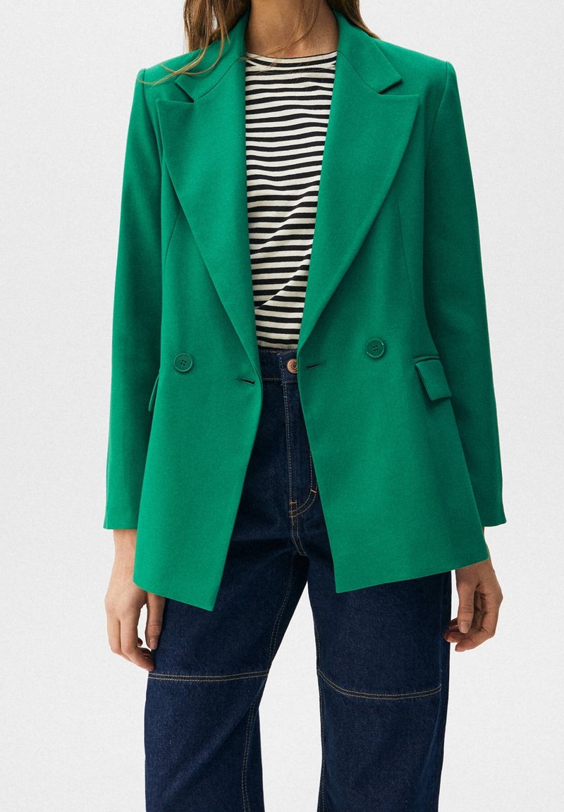 Femme portant un blazer vert à double boutonnage sur une chemise rayée noir et blanc, associée à un jean bleu foncé.