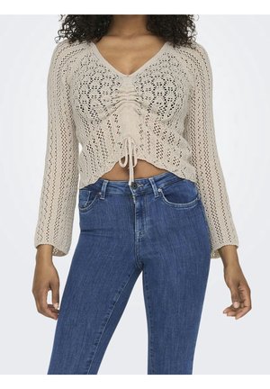 Femme portant un pull court beige en maille avec fronces sur le devant et cordon, associé à un jean bleu taille haute sur fond uni.