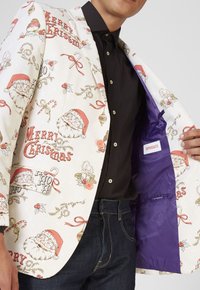 Witte blazer met een kerstthema print met daarop de kerstman, hulst en versieringen. Paarse voering en twee oranje knopen aan de voorkant.