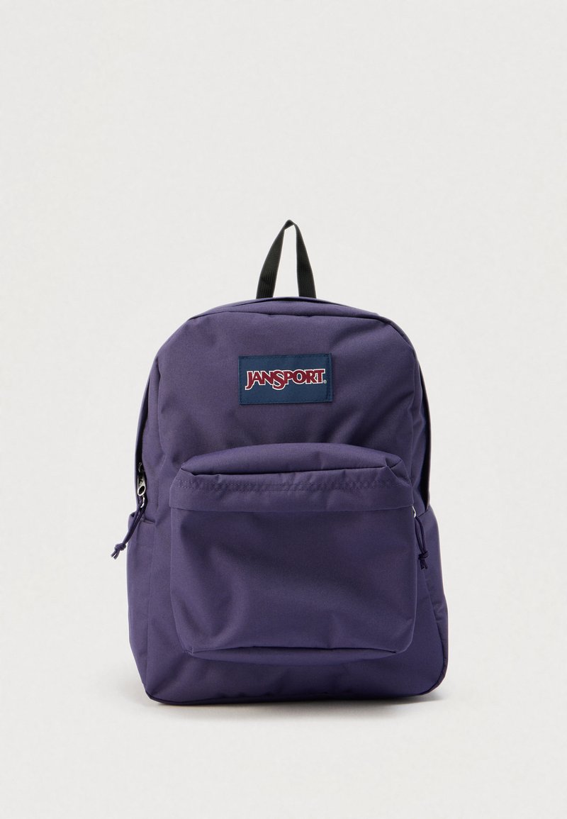 Sac à dos JanSport violet avec une poche avant zippée, une poignée noire sur le dessus, et un écusson de logo de marque visible sur un fond clair uni.