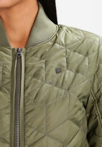 Quilted green bomber jacket with a ribbed collar, front zipper, and a small brand logo on the chest. Lightly textured fabric.