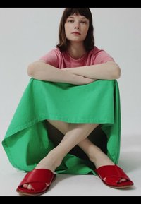 Mladá žena s hnedými vlasmi sediaca v tureckom sede, oblečená v ružovej košeli, zelenej sukni a červených šľapkách s otvorenou špičkou na jednoduchom svetlom pozadí.