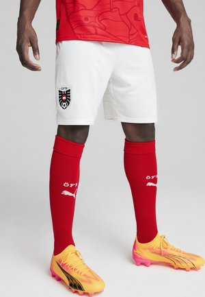 Rotes Trikot mit einem dezenten Muster, weiße Shorts mit einem Logo, rote Socken und auffällige gelbe und rosa Fußballschuhe mit schwarzen Akzenten.