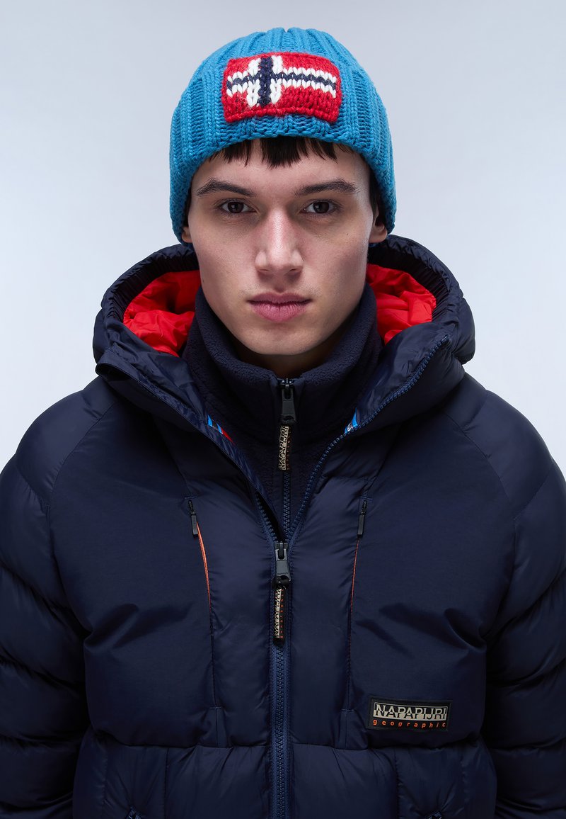 Blauer gestrickter Beanie mit rot-weißen Flaggenmuster, kombiniert mit einer marineblauen Pufferjacke mit rotem Innenfutter und Reißverschlüssen sowie einem Logo-Patch.