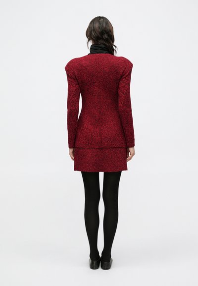 Červené šaty s vzorom, so strukturovanými ramenami, priliehavým živôtikom a viacvrstvovou sukňou, doplnené o čierne pančuchy a topánky, pohľad zozadu.