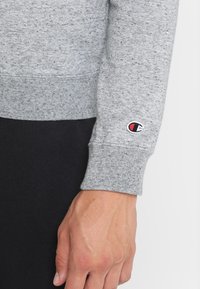 Felpa grigia con finitura testurizzata, caratterizzata da un polsino elastico nero e un piccolo logo Champion ricamato in rosso e blu sulla manica.