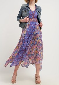 Maxi šaty v purpurové barvě s paisley a květinovými vzory, doplněné červeným vyšívaným pasem. Zkombinováno s džínovou bundou a platformovými sandály.