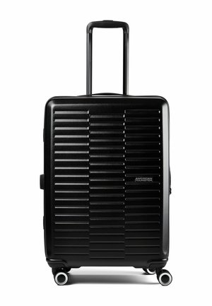 Valigia rigida nera con scanalature orizzontali, quattro ruote girevoli, manico telescopico esteso e logo "American Tourister" sulla parte anteriore.