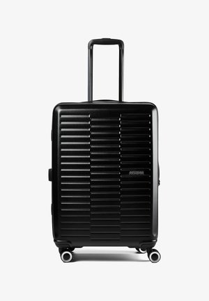 Valigia rigida nera con scanalature orizzontali, quattro ruote girevoli, manico telescopico esteso e logo "American Tourister" sulla parte anteriore.