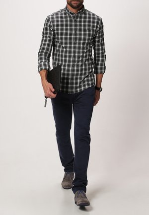Camicia a quadri bianca e nera da uomo, abbottonata, con pantaloni blu navy, che tiene una pochette nera dall'aspetto testurizzato e indossa scarpe grigie.