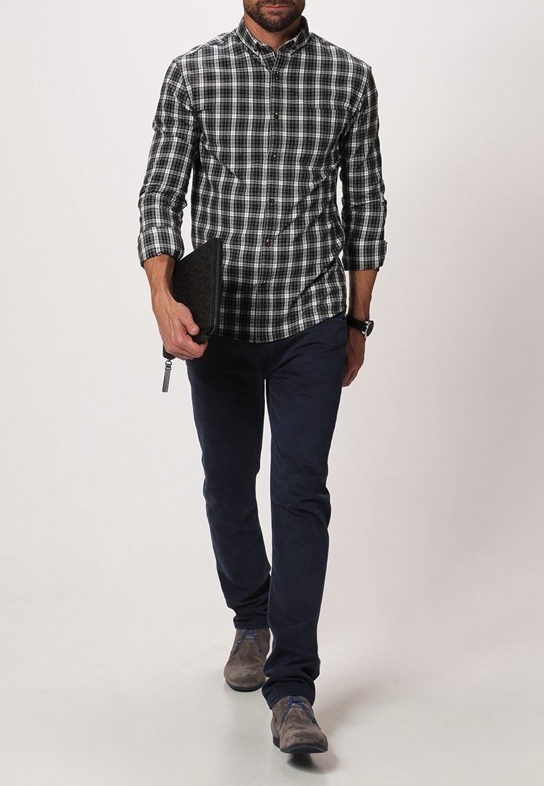 Svart och vit rutig skjorta för män, knäppt, med marinblå byxor, hållande en strukturerad svart clutch och bärande grå skor.