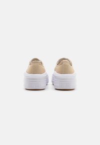 Beige canvas sneakers met een gevoerde kraag, witte rubberen zolen en een gestructureerd platform, voorzien van "Converse All Star" branding aan de achterkant.