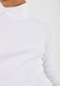 Valkoinen ribbikuvioitu poolopaita, jossa pitkät hihat. Kangas näyttää sileältä ja vartalonmyötäiseltä, ja siinä on korkea kaulus ilman näkyviä kuvioita.