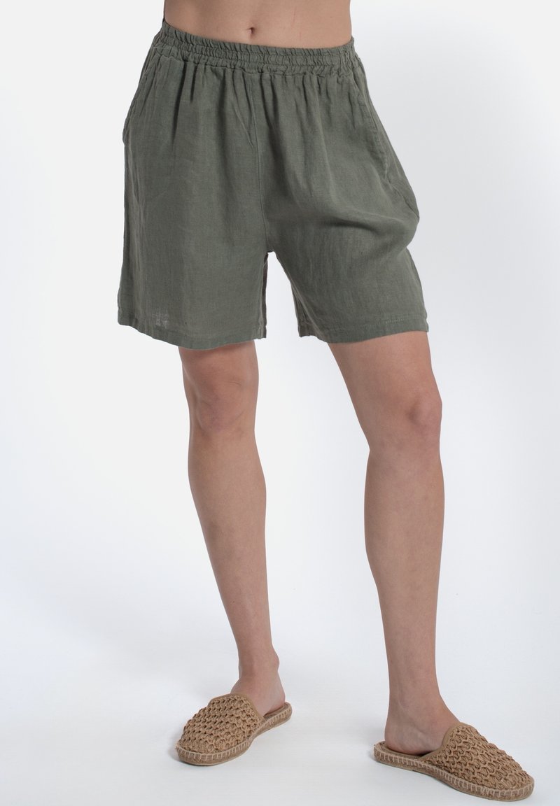 Pantaloni corti in lino verde oliva con vita elasticizzata e tasche laterali. Vestibilità comoda, taglio fino al ginocchio, tessuto testurizzato. Indossati con pantofole intrecciate.