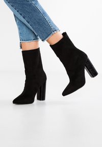 Botines de ante negros con punta afilada y tacones de bloque texturizados en negro, combinados con unos jeans cortos de mezclilla azul desgastados.