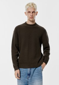 Pulover tricotat cu nervuri de un verde închis, cu guler înalt și o croială relaxată, asortat cu blugi din denim de culoare albastru deschis.