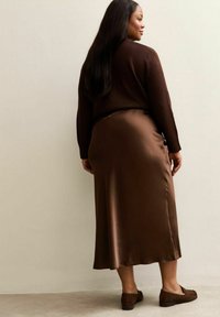 Donkerbruine trui gecombineerd met een lange, satijnen, bruine rok, voorzien van een gladde textuur en subtiele glans. Compleet met bruine instappers.