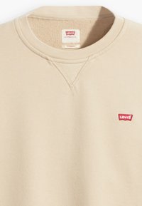 Beige sweater gemaakt van zacht materiaal, met een ronde hals, ribgebreide mouwen en een klein rood Levi's logo op de linkerkant van de borst. De textuur is glad.