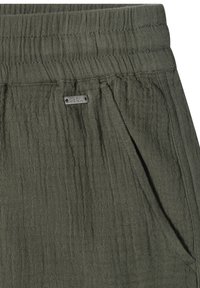 Pantalon en tissu texturé vert olive avec taille élastique, poche latérale et petite étiquette métallique rectangulaire de marque près de la poche.