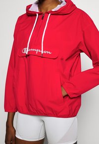 Person klädd i en röd Champion-hoodie med vita snören och vita shorts, med handen i framfickan, stående mot en enfärgad bakgrund.