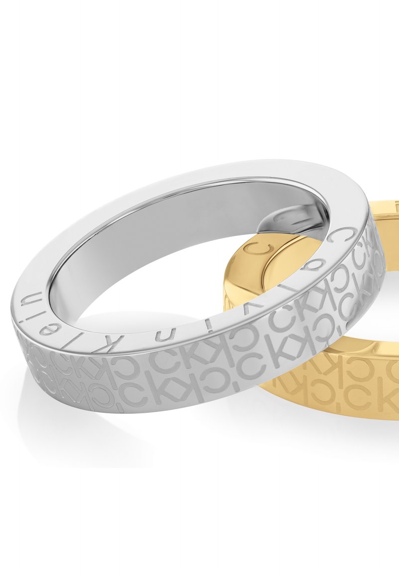Deux anneaux : l'un en argent avec une finition lisse et gravé d'un motif "CK", l'autre en or avec une texture mate et la même gravure.