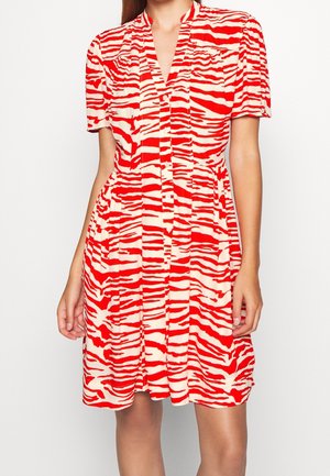 Mujer con un vestido abotonado de manga corta con estampado de cebra en rojo y blanco y una falda ligeramente plisada.