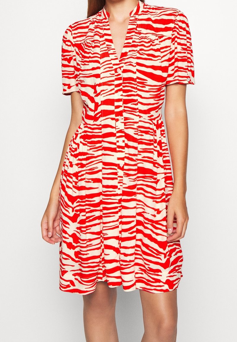 Femme portant une robe boutonnée à manches courtes avec un imprimé zèbre rouge et blanc et une jupe légèrement plissée.