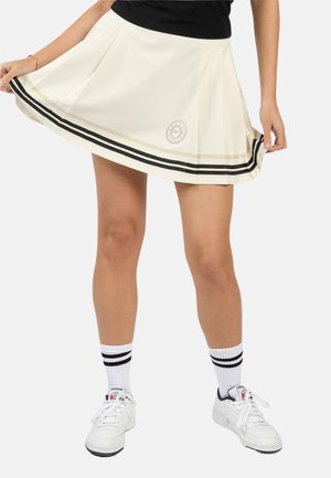 Frau trägt einen weißen plissierten Rock mit schwarzen und beigen Streifen am Saum, kombiniert mit weißen Sneakers und gestreiften Socken.