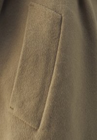 Detail vlneného kabáta s nášivkou; jemná, plstená textúra; teplá béžová farba; minimalistický dizajn s čistými líniami a bez viditeľných kovových častí.