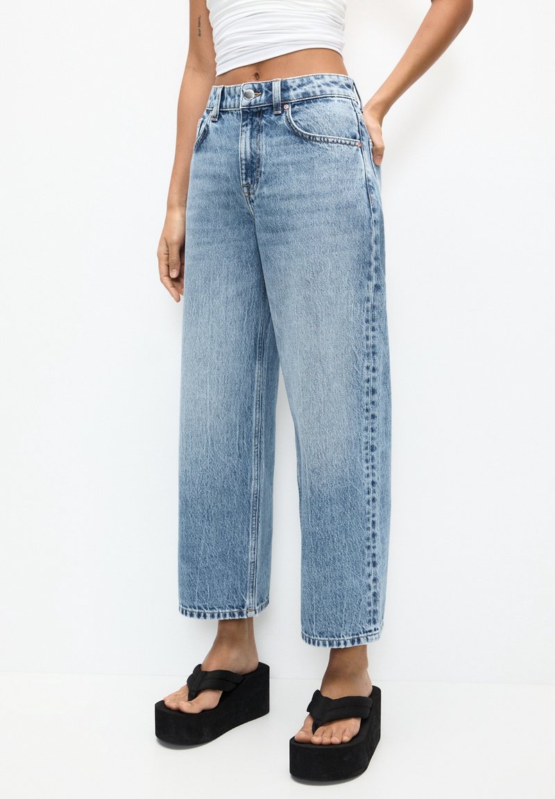 Jean bleu à jambes larges en denim, avec une taille haute, une texture délavée et des surpiqûres contrastées. Porté avec des sandales à plateforme noires.