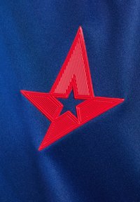 Röd gummistjärna logotyp på en blå tygbakgrund; logotypen har upphöjda linjer och en stjärnformad urtagning i mitten. Strukturerat material.