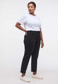 Egy nő magabiztosan áll, fehér pólót, fekete, magas derekú farmernadrágot és fehér sportcipőt visel, kezét a csípőjén tartva.