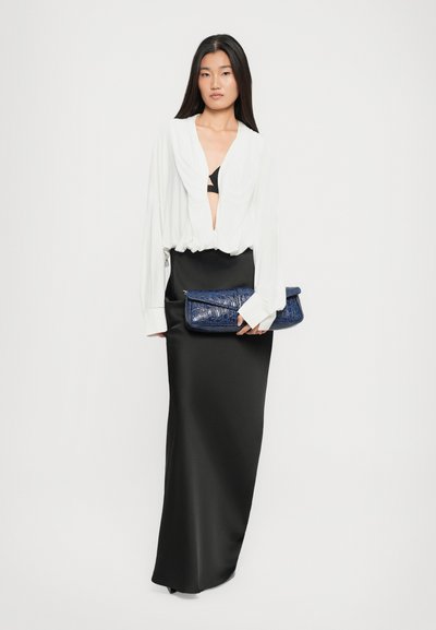 Donna che indossa una camicia bianca con scollo a V profondo e una lunga gonna nera, che tiene una pochette blu scuro testurizzata su uno sfondo bianco semplice.