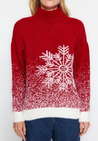 Punainen neulepaita, jossa on korkea kaulus, valkoisia lumihiutaleita esittävää kuviota ja väriharjoitusta, pehmeä pinta ja valkoiset ribbiresorit.