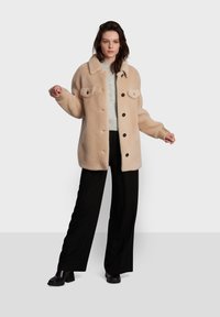 Casaco de pelo sintético bege com decote collared, bolsos frontais e fecho com botão, combinado com um suéter cinza e calças largas pretas.