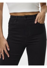 Femme portant un jean noir taille haute et un top court blanc, une main dans la poche et l'autre reposant sur la cuisse.