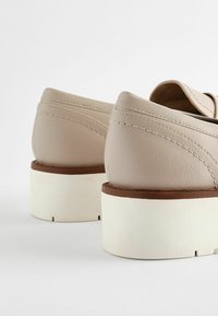 Beige tekonahkakengät pyöreillä taustoilla, joissa on valkoiset platform-pohjat ja ruskeat yksityiskohdat. Reunoja koristaa ommeltu yksityiskohta.