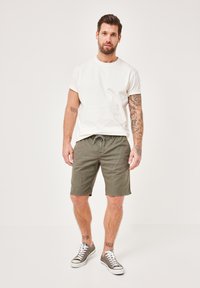 Weiße Baumwoll-T-Shirt, olivgrüne Shorts mit Kordelzugbund und grauen Sneakers. Sichtbare Tattoos an den Knöcheln, stehend vor einem neutralen Hintergrund.